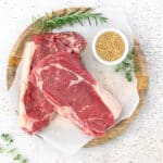 Rauwe ribeye steaks op een houten bord met rozemarijn, tijm en mosterdzaadjes.