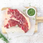 Een rauwe T-bone steak op een houten plank met verse kruiden en rozemarijn erbij.